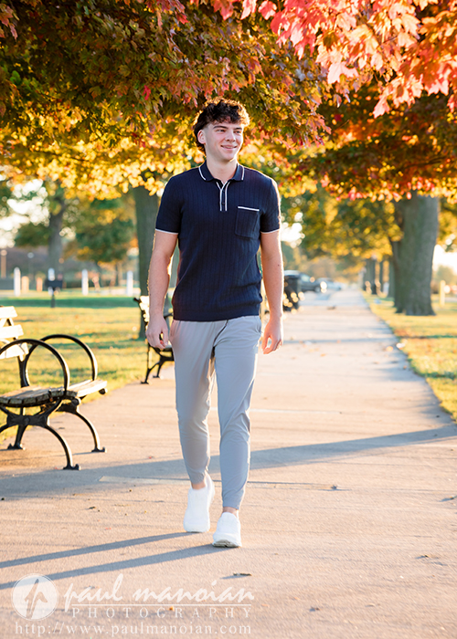 A young man in light pants and a dark shirt walks along a sunlit park path lined with benches and colorful autumn trees during his Detroit senior pictures session.