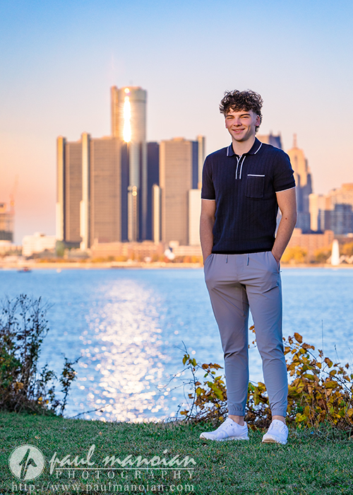 A young man stands smiling on grass by a river at sunset, with a city skyline and tall buildings in the background—capturing his Detroit senior pictures session. He is wearing a dark polo shirt, light pants, and white sneakers. Logo and website are at the bottom.