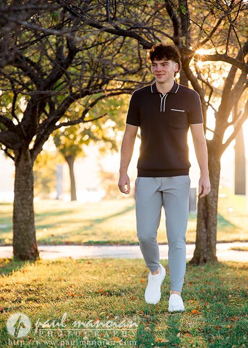 A young man in a dark polo shirt and light pants walks on grass under trees at sunset, smiling. During his Detroit senior pictures session, sunlight filters through the branches, creating a warm and peaceful atmosphere.