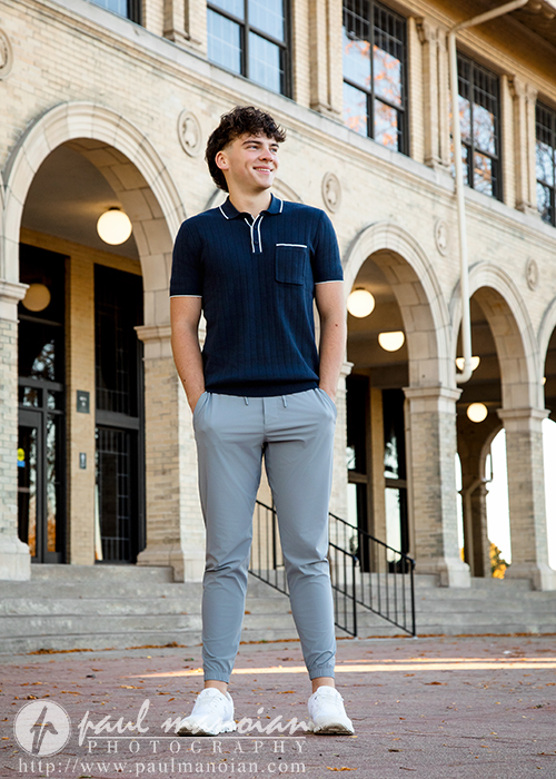 A young man stands smiling in front of a brick building with arched windows during his Detroit senior pictures session, wearing a navy polo shirt, light gray pants, and white sneakers. Steps and black railings lead up to the entrance behind him.