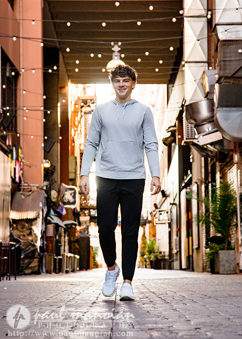 A young man with curly hair, wearing a light gray hoodie, black pants, and white sneakers, walks confidently down a cobblestone alleyway decorated with hanging string lights during his Detroit senior pictures session.