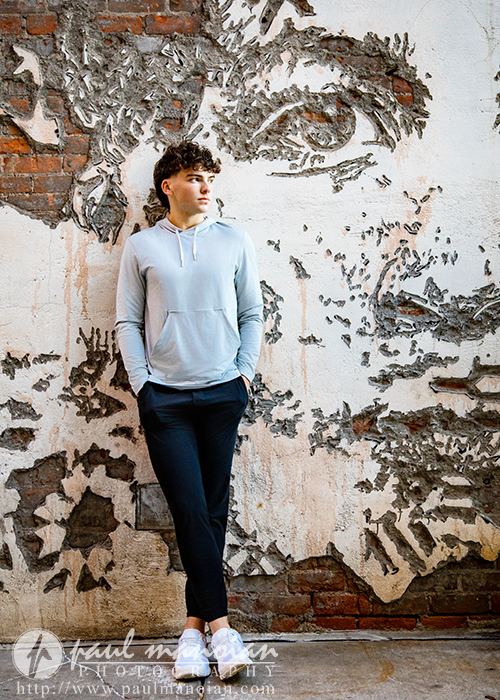 A young man in a light hoodie and dark pants stands against a brick wall with an artistic mural of a face during his Detroit senior pictures session. He has his hands in his pockets, looks to the side, and wears white shoes for a casual vibe.