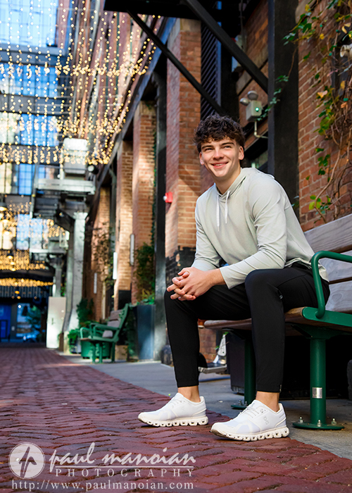 A young man with short brown hair, wearing a light hoodie, black pants, and white sneakers, sits smiling on a bench in a brick alley decorated with string lights during his Detroit senior pictures session. He is relaxed, looking toward the camera.
