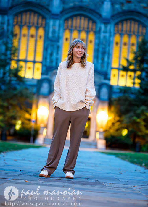 A high school senior stands on a paved path wearing a cream sweater and brown pants, smiling during her Divine Child senior pictures session. Behind her is a lit building with large arched windows, surrounded by trees at dusk.