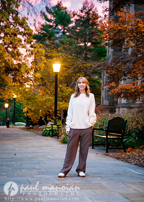 A high school senior girl in a cream sweater and brown pants stands smiling on a stone walkway lined with benches and trees at sunset, the street lamps glowing softly during her Divine Child senior pictures session.