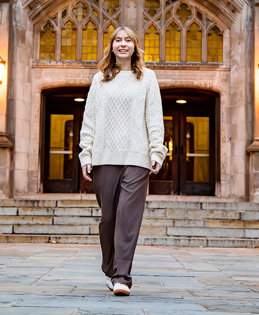 A woman in a cream sweater and brown pants walks confidently on a stone path in front of a grand building with arched doors and glowing lamps, surrounded by greenery—capturing the elegance of a Divine Child senior pictures session.