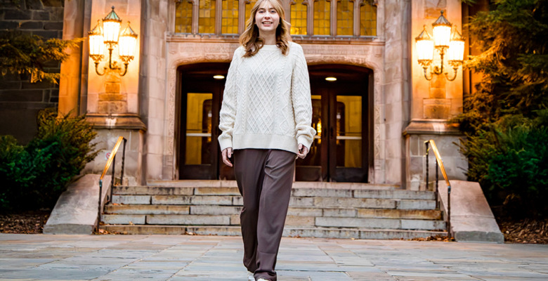 A woman in a cream sweater and brown pants walks confidently on a stone path in front of a grand building with arched doors and glowing lamps, surrounded by greenery—capturing the elegance of a Divine Child senior pictures session.