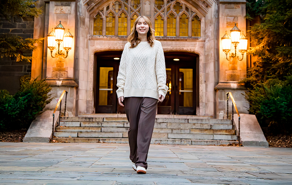 A woman in a cream sweater and brown pants walks confidently on a stone path in front of a grand building with arched doors and glowing lamps, surrounded by greenery—capturing the elegance of a Divine Child senior pictures session.