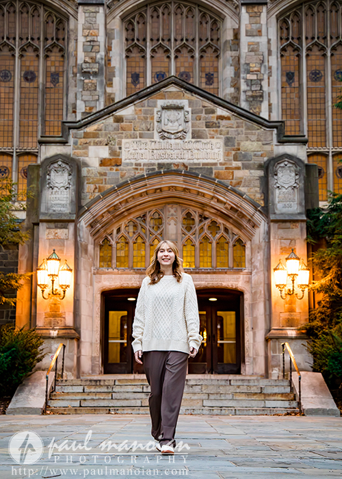 A high school senior girl in a cream sweater and brown pants stands smiling on stone steps during her Divine Child senior pictures session in front of an ornate Gothic-style university building with large arched doors and lit lanterns.