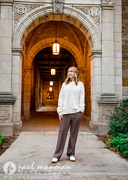 A high school senior girl wearing a cream sweater and brown pants stands smiling under a stone archway with hanging lanterns, framed by historic architecture and greenery during her Divine Child senior pictures session.