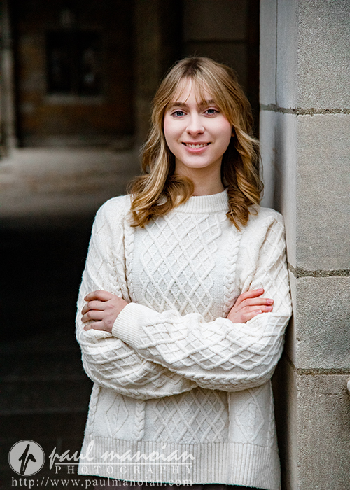 A high school senior girl with blonde hair wearing a white knitted sweater stands with arms crossed, leaning against a stone wall and smiling during her Divine Child senior pictures session. The softly blurred background suggests an outdoor or architectural setting.