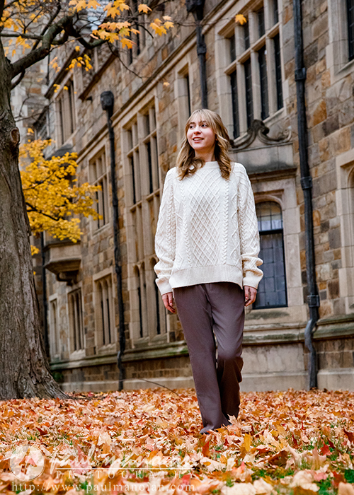 A high school senior girl wearing a cream sweater and brown pants walks through fallen autumn leaves on a sidewalk next to a historic stone building—an ideal spot for a Divine Child senior pictures session among yellow-orange trees.