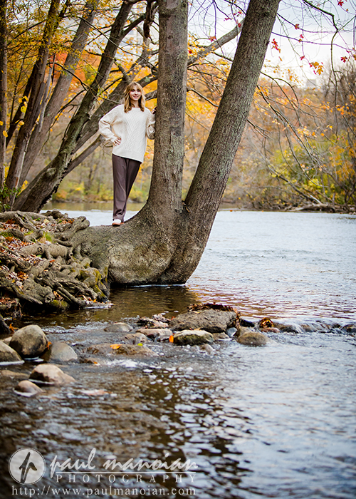 A high school senior girl in a white sweater stands on the trunk of a tree leaning over a river, surrounded by autumn foliage—an inspiring scene for a Divine Child senior pictures session. Rocks and water are in the foreground, with fall colors and trees in the background.