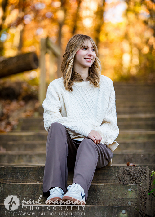 A high school senior girl with blonde hair sits on outdoor wooden steps, smiling and looking to the side during her Divine Child senior pictures session. She wears a white knitted sweater, gray pants, and white sneakers. Autumn trees with golden leaves fill the background.