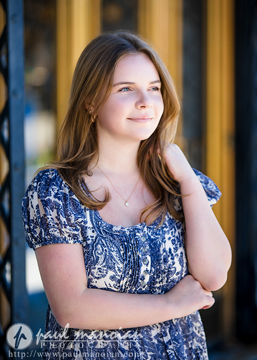 A high school senior with light brown hair stands in front of a yellow and black doorway, smiling softly. She wears a blue and white patterned dress—capturing the perfect moment during her Grosse Pointe senior pictures session.