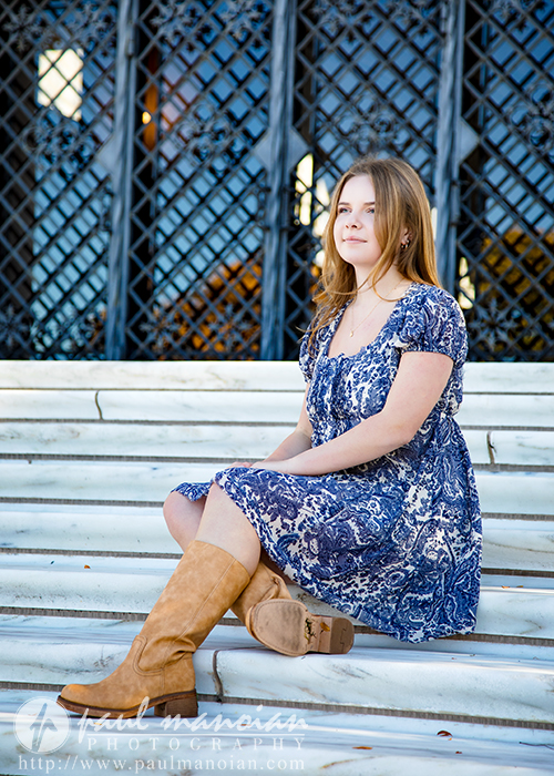 A high school senior in a blue patterned dress and tan boots sits on white marble steps in front of ornate black iron doors, looking off to the side with a calm expression during her Grosse Pointe senior pictures session.