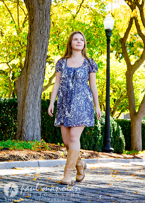A high school senior in a blue patterned dress and tan boots walks confidently on a cobblestone path lined with trees and greenery under bright sunlight during her Grosse Pointe senior pictures session.