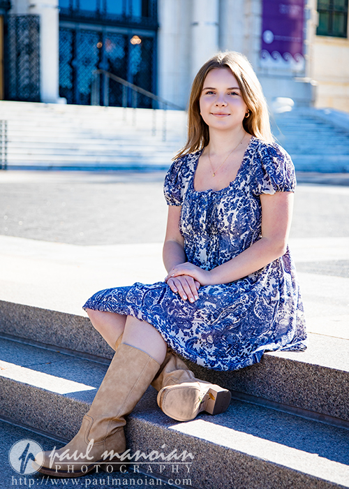 A high school senior with long hair sits on outdoor stone steps, wearing a blue patterned dress and tan knee-high boots. She smiles softly, hands resting on her knee, during her Grosse Pointe senior pictures session. A building entrance is in the background.