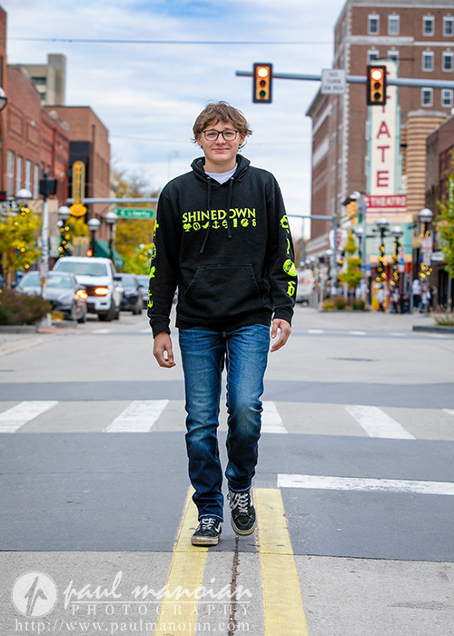 A teenage boy wearing glasses, a black "Shinedown" hoodie, and jeans walks confidently across a city street crosswalk during his Jackson High School senior pictures session, with cars, buildings, and traffic lights in the background.