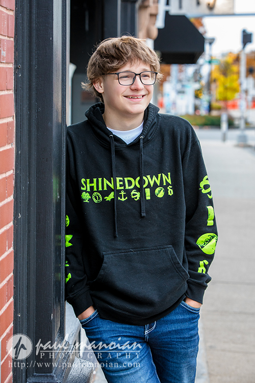 A smiling teenager with glasses, wearing a black Shinedown hoodie and jeans, stands on a city sidewalk with hands in pockets, leaning against a brick wall during a Jackson High School senior pictures session. Urban street and autumn trees fill the background.