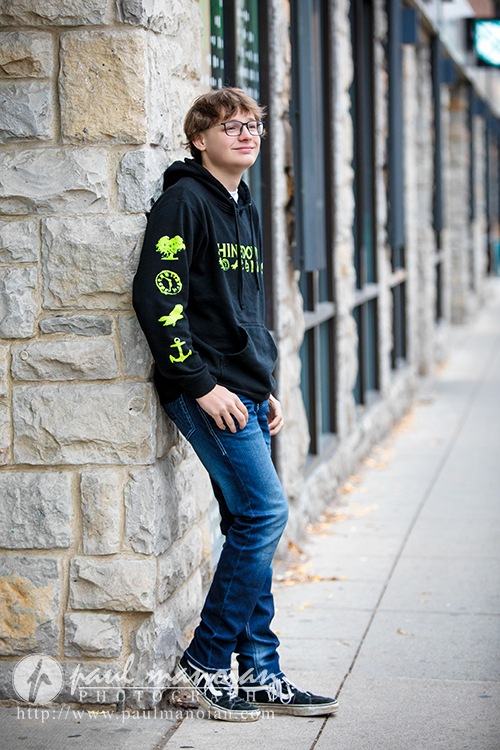 Teenage boy with glasses, wearing a black hoodie and blue jeans, leans casually against a stone wall on a sidewalk, smiling slightly during his Jackson High School senior pictures session. The background features windows and a street scene.