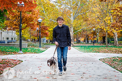 A Jackson High School senior walks a brown and white dog on a leaf-covered path in a park with colorful autumn trees during his senior pictures session.