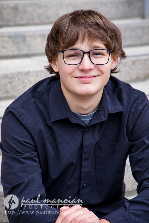 A young person with light skin, brown hair, and glasses sits on outdoor stone steps, smiling and wearing a navy button-up shirt over a gray t-shirt during their Jackson High School senior pictures session. The logo "paul mancan photographer" is visible in the corner.