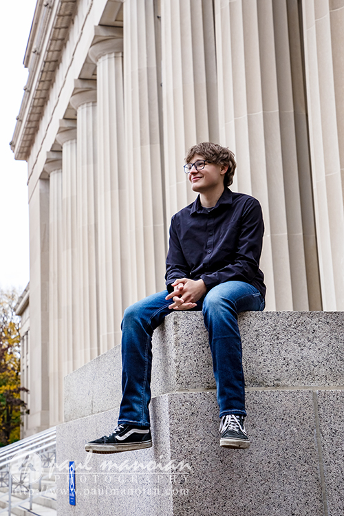 A young person with glasses, wearing a dark shirt and blue jeans, sits smiling on a stone ledge in front of a large building with tall columns during their Jackson High School senior pictures session.