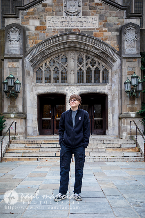 A person wearing glasses, a black jacket, and dark pants stands on stone steps in front of a large arched entrance to an ornate stone building—an ideal spot for a Jackson High School senior pictures session.