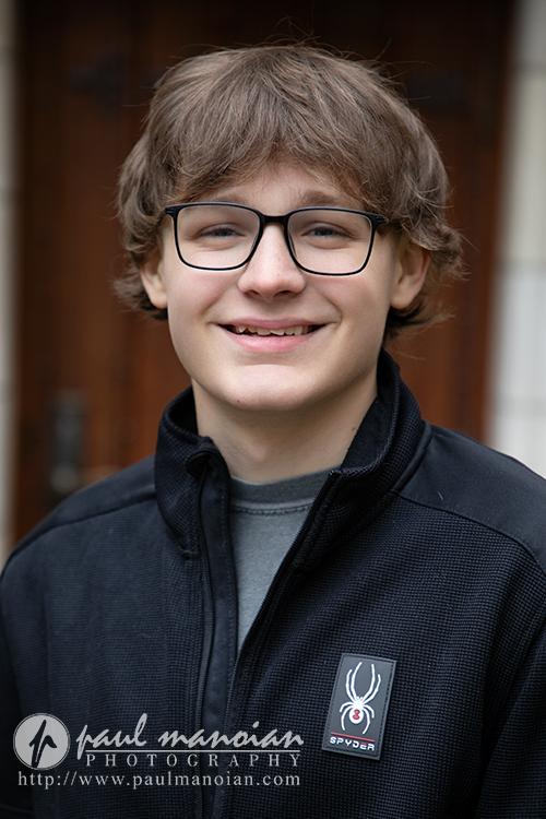 A teenage boy with glasses and medium-length brown hair smiles at the camera during his Jackson High School senior pictures session. He is wearing a black jacket with a Spider logo patch; a wooden door is slightly blurred in the background.
