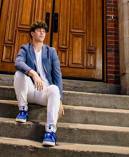 A young man with curly hair sits on outdoor stone steps in front of large wooden doors, dressed in a blue blazer, white pants, and blue sneakers, looking to the side during his Macomb senior pictures session.