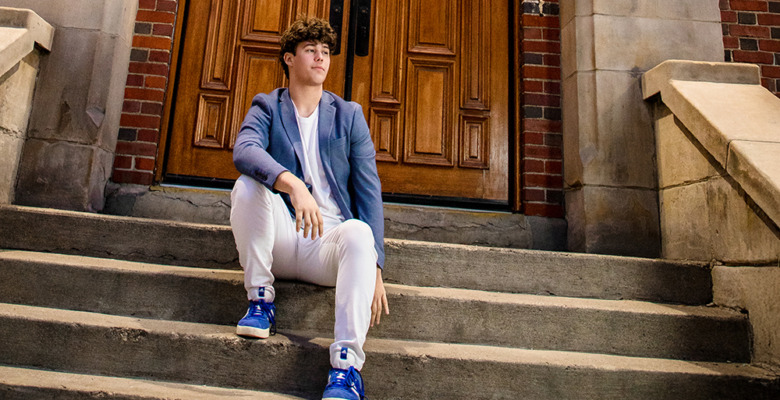 A young man with curly hair sits on outdoor stone steps in front of large wooden doors, dressed in a blue blazer, white pants, and blue sneakers, looking to the side during his Macomb senior pictures session.