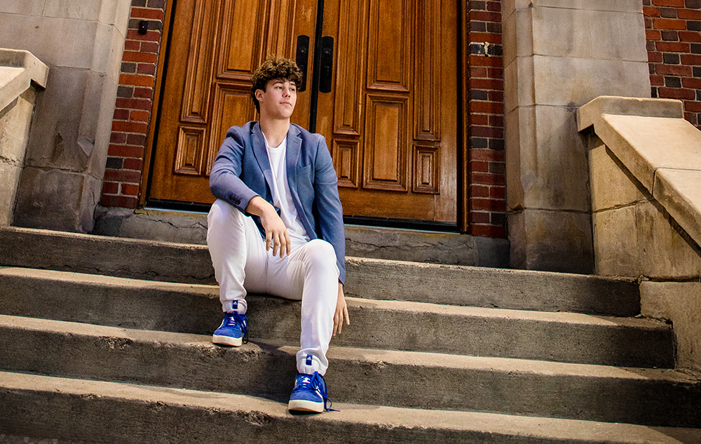 A young man with curly hair sits on outdoor stone steps in front of large wooden doors, dressed in a blue blazer, white pants, and blue sneakers, looking to the side during his Macomb senior pictures session.