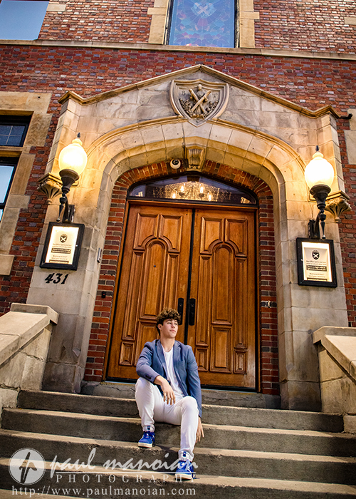 A high school senior in a blue jacket and white pants sits on the steps outside a brick building with large wooden double doors—an inviting spot for a Macomb senior pictures session, complete with two lit lanterns and plaques flanking the entrance.