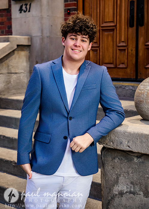 A young man with curly brown hair, wearing a blue blazer and white shirt, stands and smiles in front of stone steps and a wooden door during his Macomb senior pictures session.