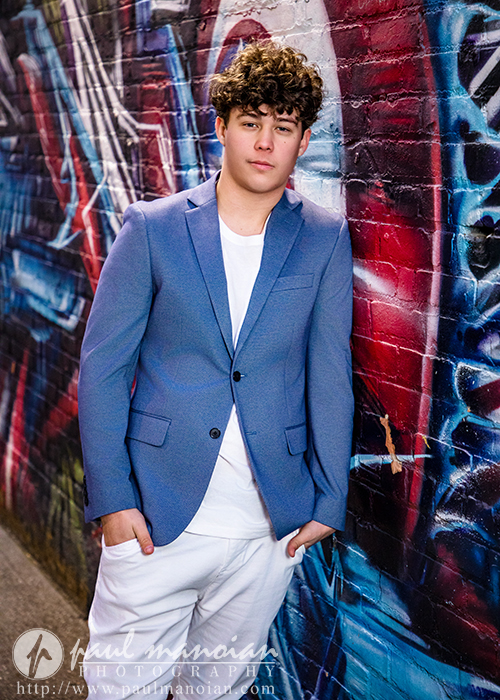 A high school senior with curly hair in a blue blazer and white pants leans confidently against a colorful graffiti-covered brick wall, capturing the vibrant spirit of his Macomb senior pictures session.