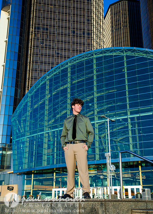 A young man in a green dress shirt and khaki pants stands confidently on concrete steps in front of a modern glass building, captured during his Macomb senior pictures session with tall skyscrapers rising in the clear blue sky behind him.