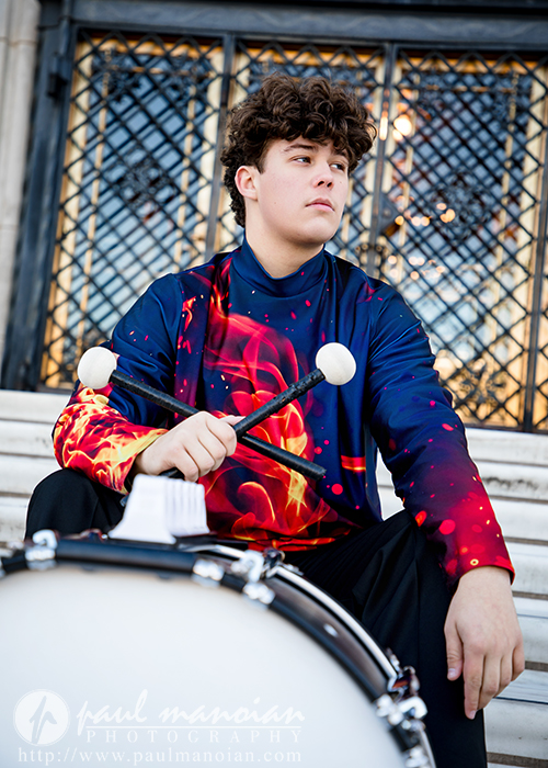 A high school senior in a colorful, fiery-patterned marching band uniform sits on outdoor steps, holding drum mallets and resting them on a snare drum during their Macomb senior pictures session, with a decorative metal gate in the background.