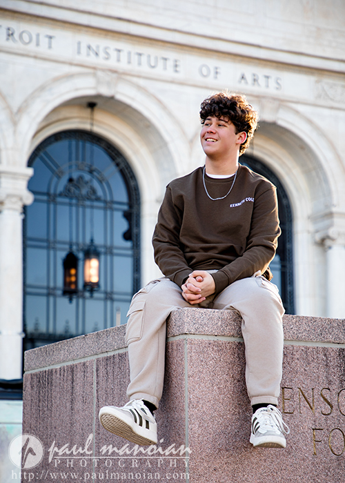 A high school senior with curly hair, wearing a brown sweatshirt and light pants, smiles and looks to the side while sitting on a stone monument in front of the Detroit Institute of Arts during his Macomb senior pictures session.
