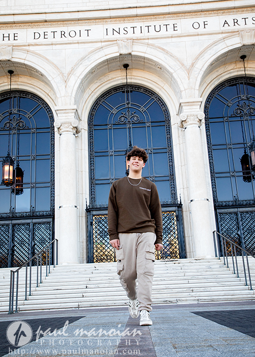 A high school senior in casual clothes walks down the steps outside the Detroit Institute of Arts, with large arched doorways and ornate lanterns behind him during his Macomb senior pictures session.