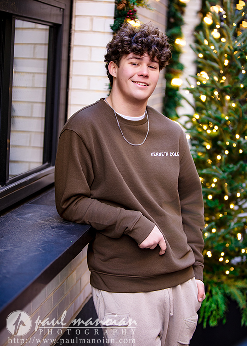 A high school senior with curly brown hair, wearing a brown Kenneth Cole sweatshirt and beige pants, leans against a black window ledge, smiling during their Macomb senior pictures session. A decorated, lit Christmas tree glows in the background.