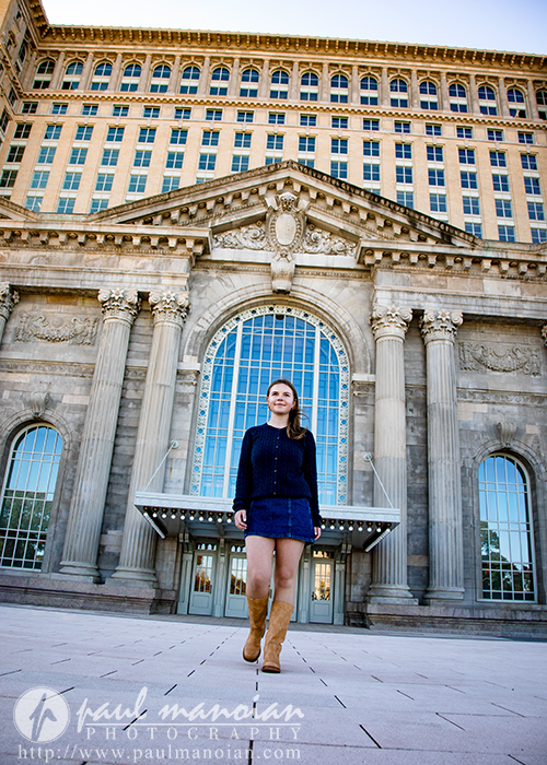 A high school senior wearing a navy sweater and boots stands smiling in front of a large historic building with tall columns and arched windows during her Grosse Pointe senior pictures session. The photo is taken from a low angle.