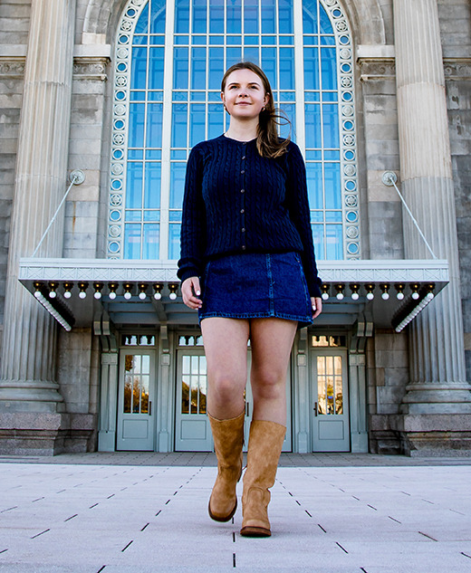 A young woman wearing a dark sweater, denim skirt, and tan boots walks confidently on a paved area in front of a grand building—an ideal location for a Grosse Pointe senior pictures session.