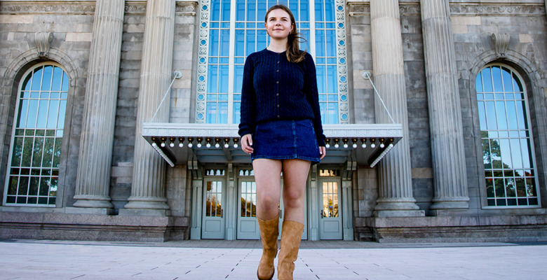 A young woman wearing a dark sweater, denim skirt, and tan boots walks confidently on a paved area in front of a grand building—an ideal location for a Grosse Pointe senior pictures session.