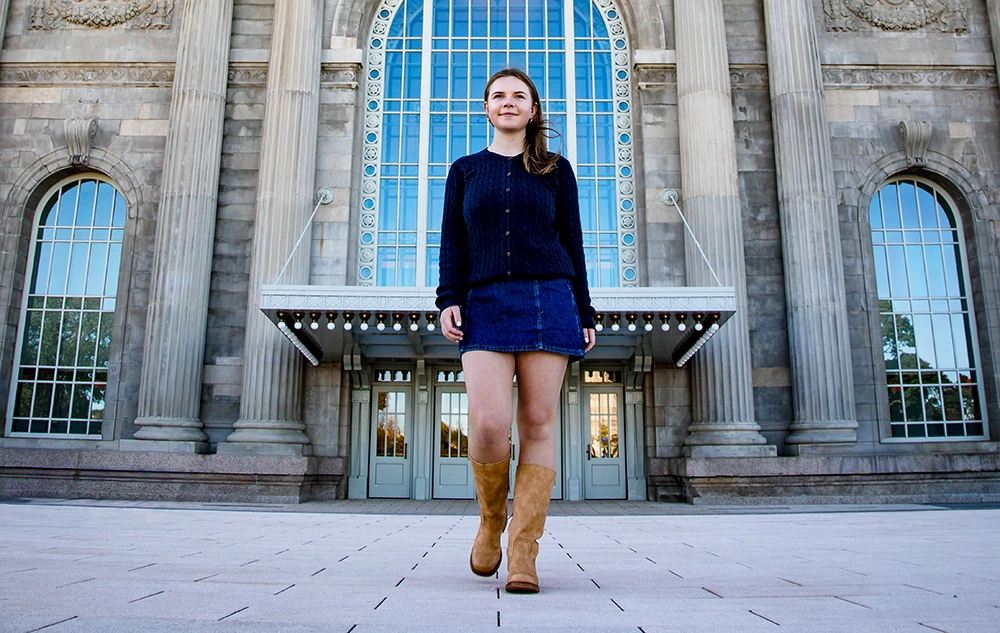 A young woman wearing a dark sweater, denim skirt, and tan boots walks confidently on a paved area in front of a grand building—an ideal location for a Grosse Pointe senior pictures session.