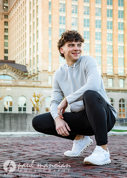 A young man with curly hair, wearing a light gray hoodie, black pants, and white sneakers, crouches and smiles outdoors on a brick path with tall buildings in the background during his Detroit senior pictures session.