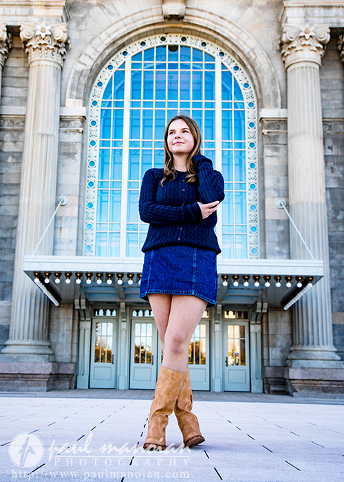 A high school senior in a blue sweater and denim skirt stands confidently with crossed arms and brown boots in front of a grand building—an ideal scene for a Grosse Pointe senior pictures session.