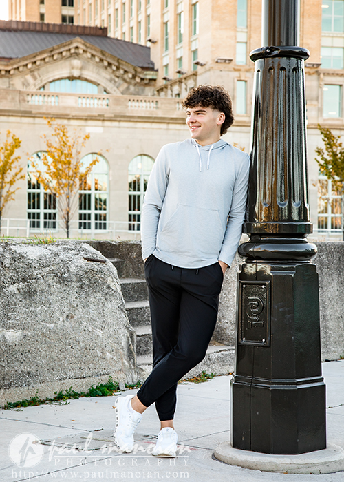 A young man in a light gray hoodie and black pants leans casually against a black lamp post outdoors, smiling during his Detroit senior pictures session. A large building with arched windows and autumn trees is in the background.