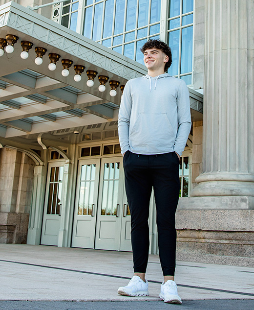 A young man in a light hoodie, black pants, and white sneakers stands smiling in front of a grand building with tall columns and large glass windows during his Detroit senior pictures session.