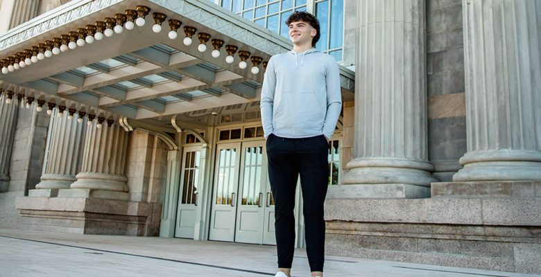 A young man in a light hoodie, black pants, and white sneakers stands smiling in front of a grand building with tall columns and large glass windows during his Detroit senior pictures session.